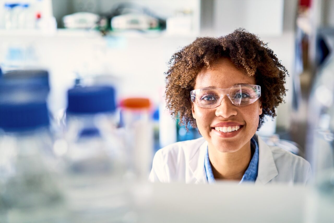 Portrait of a smiling young female scientist looking through shelves full of sample bottle while working in a lab