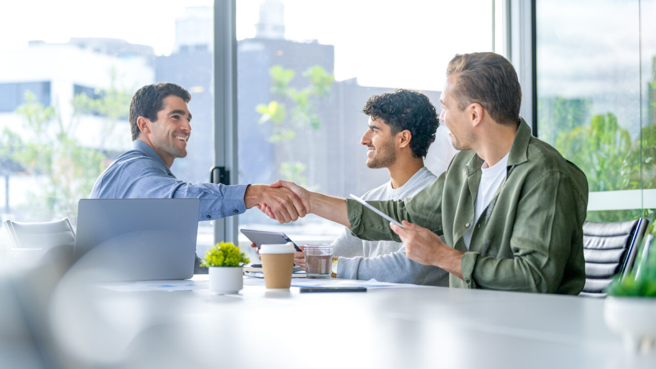 Three business men meeting with a handshake in the board room. They are using computers and digital tablets. There is a large window behind them