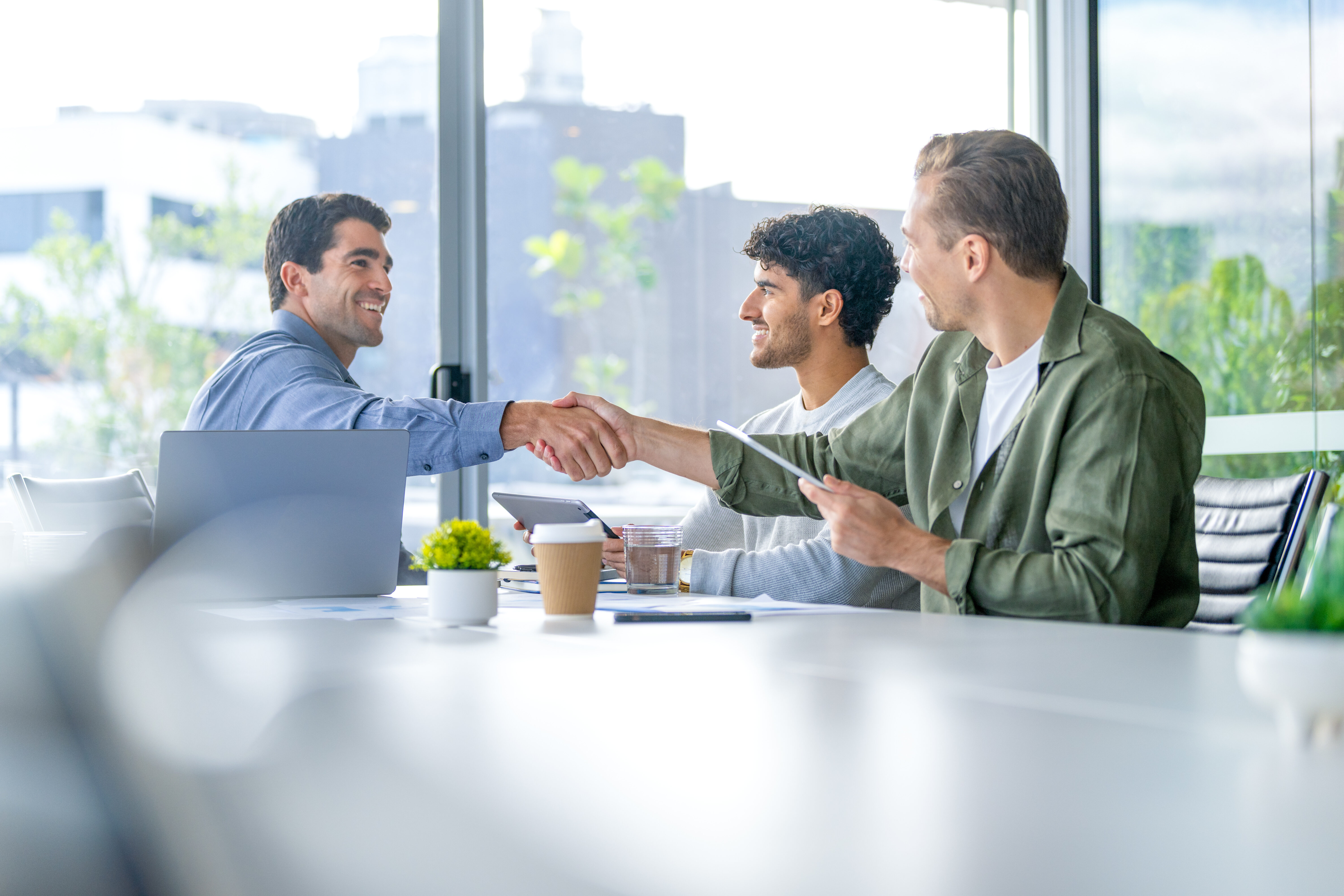 Three business men meeting with a handshake in the board room. They are using computers and digital tablets. There is a large window behind them