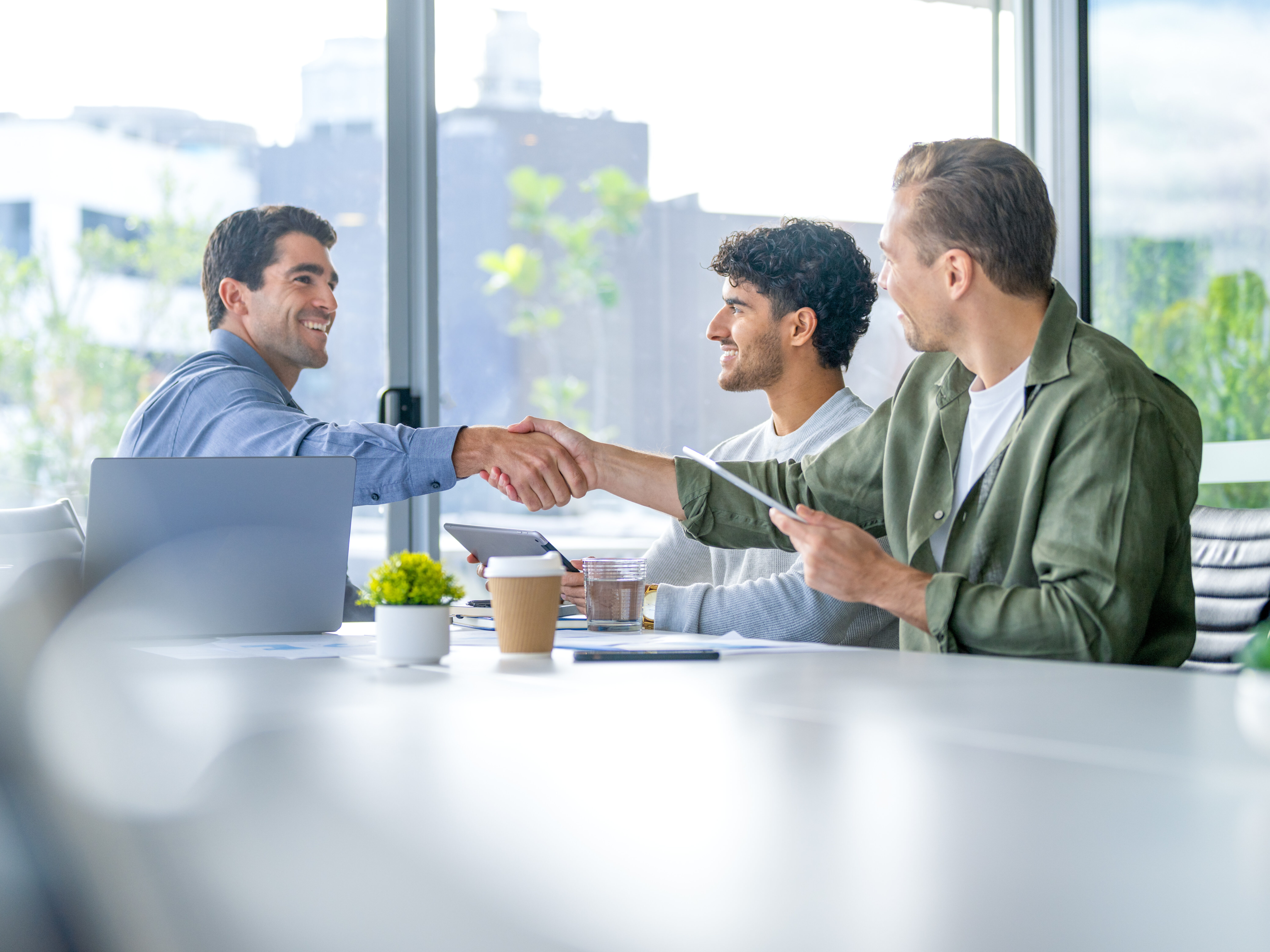 Three business men meeting with a handshake in the board room. They are using computers and digital tablets. There is a large window behind them